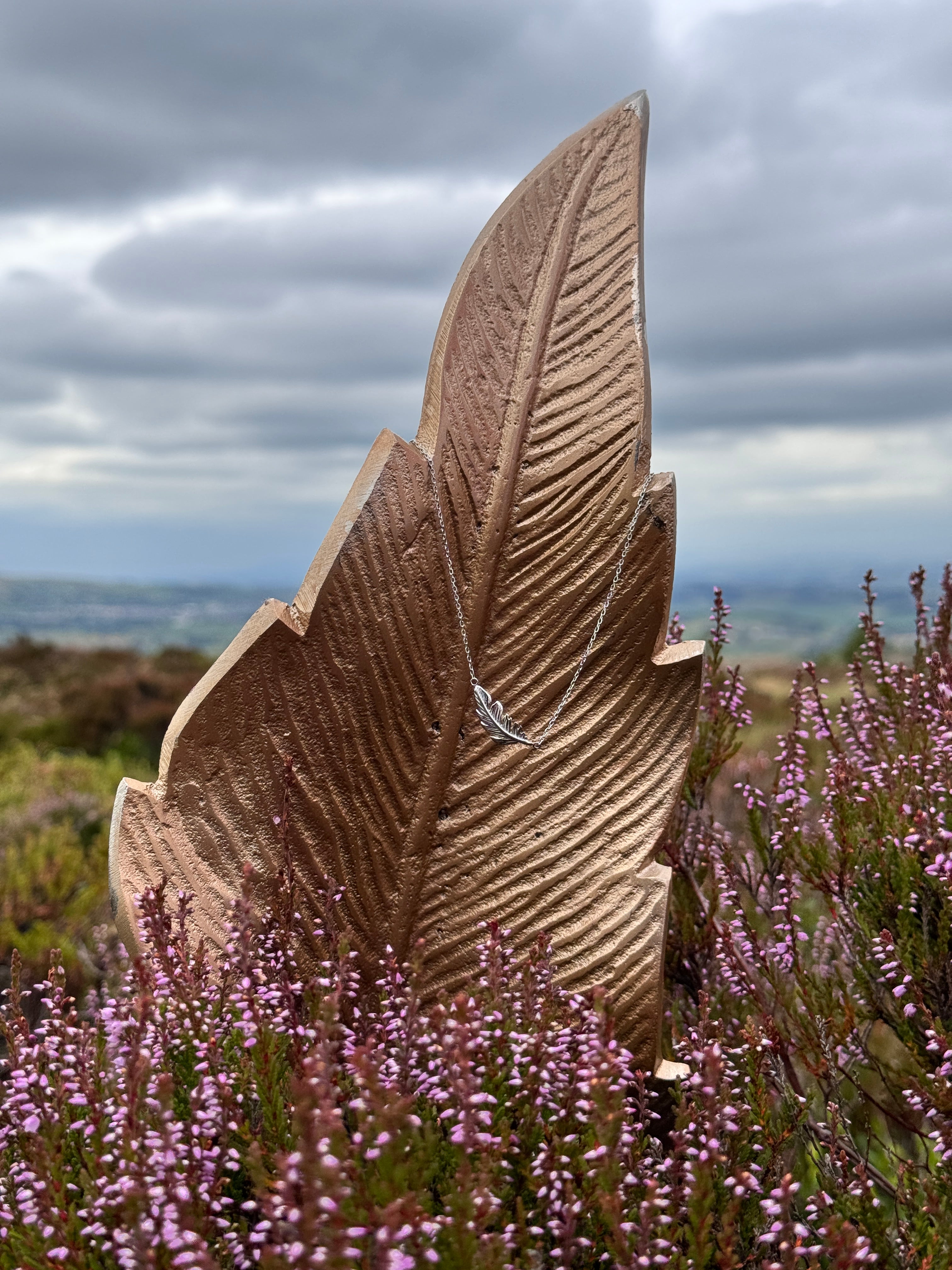 A large, textured brown feather sculpture or garden feature displayed outdoors in a field of purple heather under a cloudy sky.