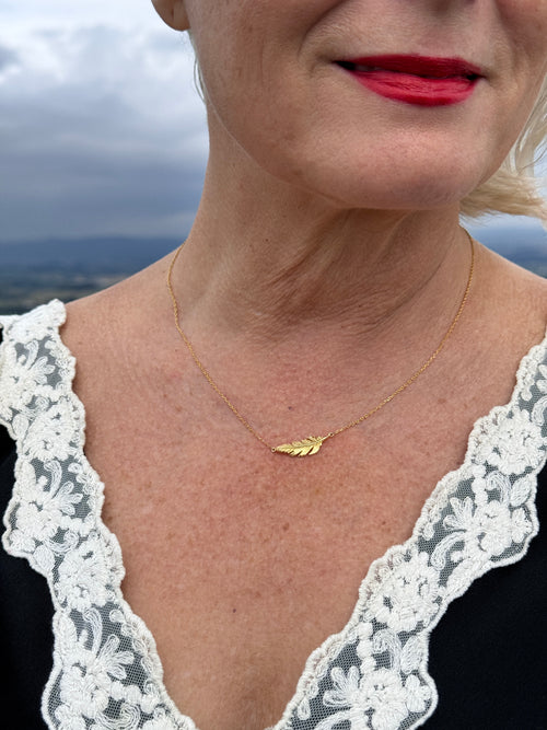 A close-up lifestyle shot of a woman wearing a delicate gold feather pendant necklace over a white lace top.