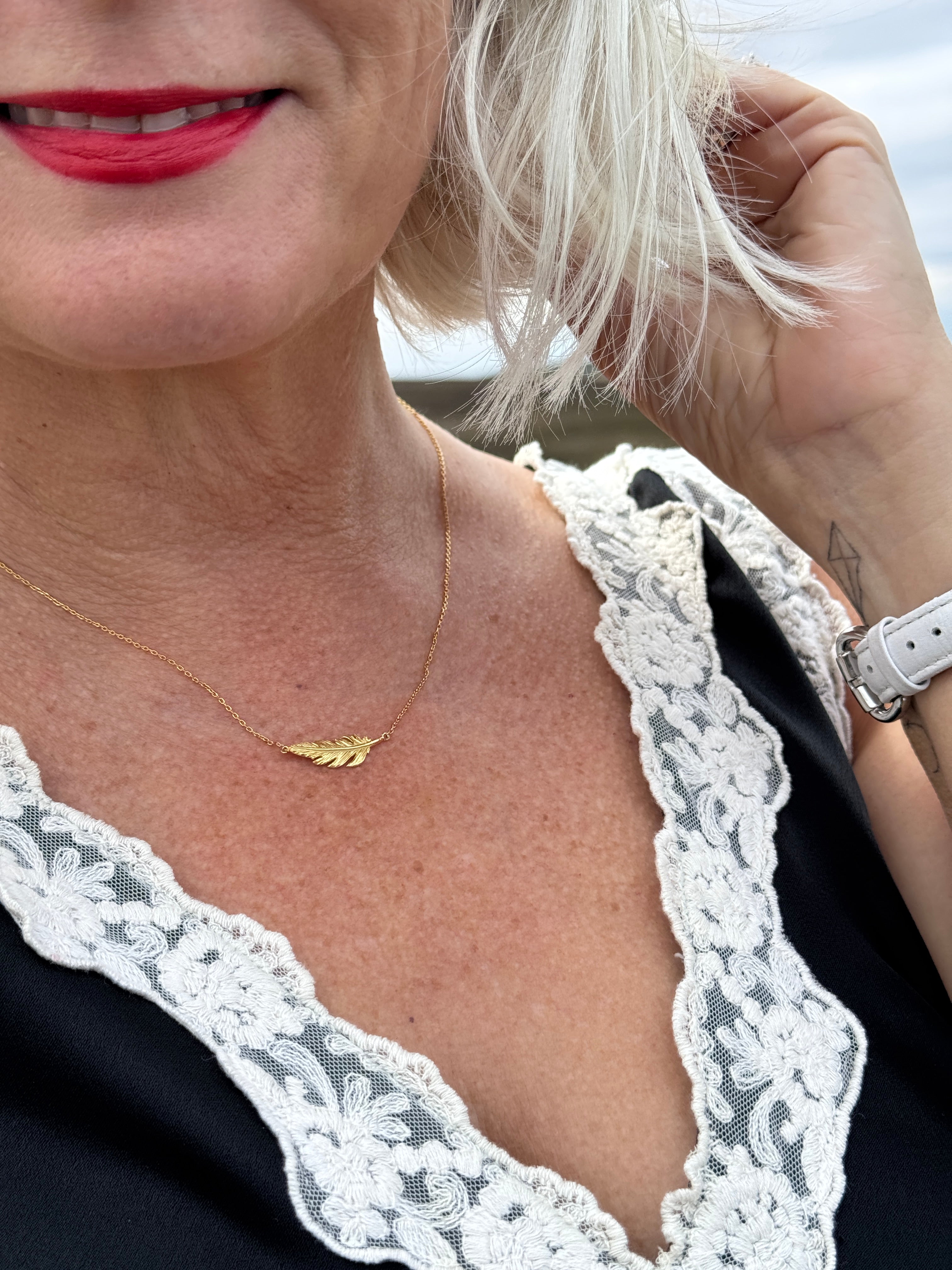 A close-up lifestyle shot of a person wearing a delicate gold necklace with a small feather pendant, styled with a black top and white lace trim.