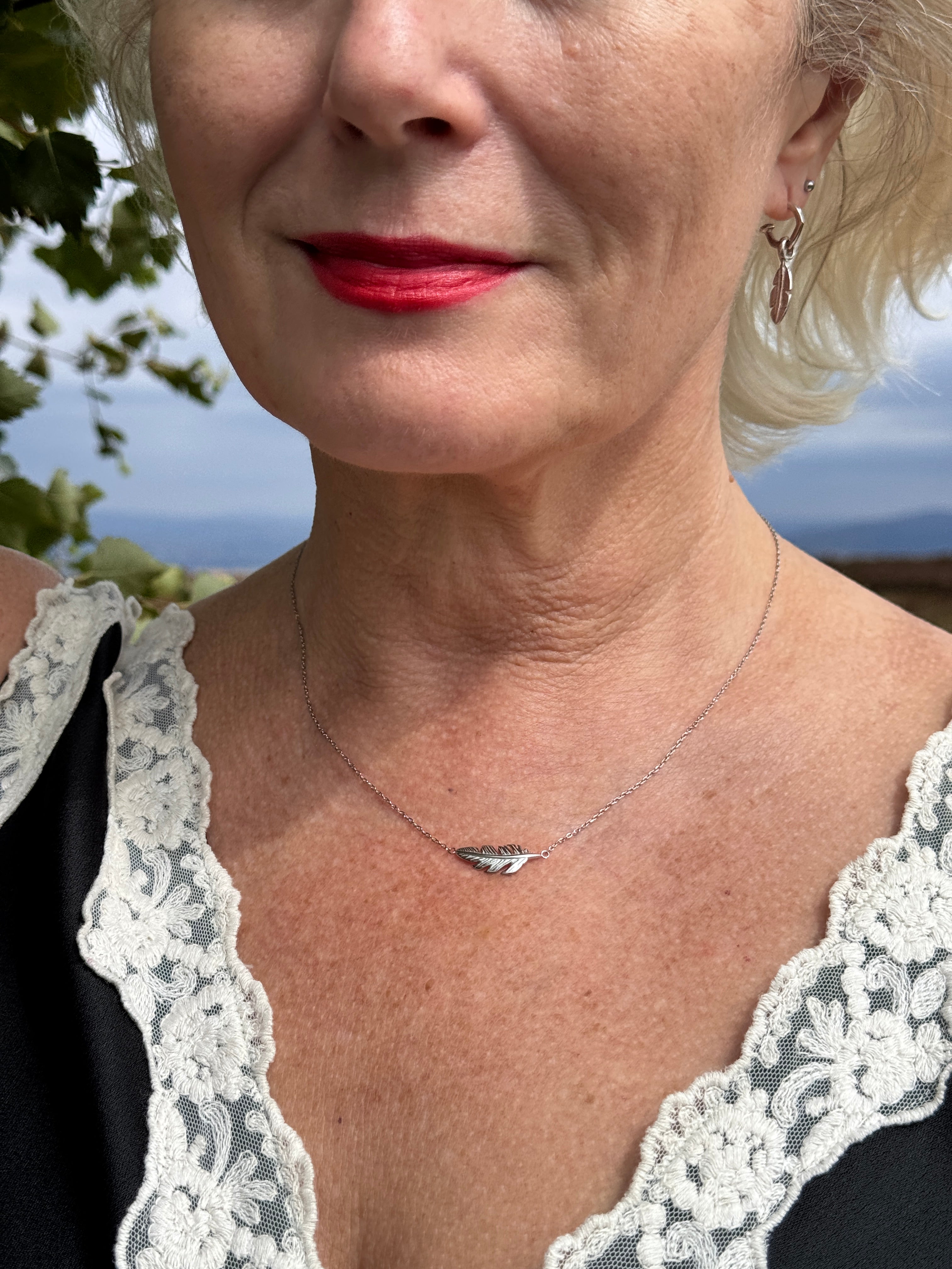 A close-up lifestyle shot of a woman wearing a delicate silver necklace with a small feather shaped pendant, styled with a black top featuring white lace trim.