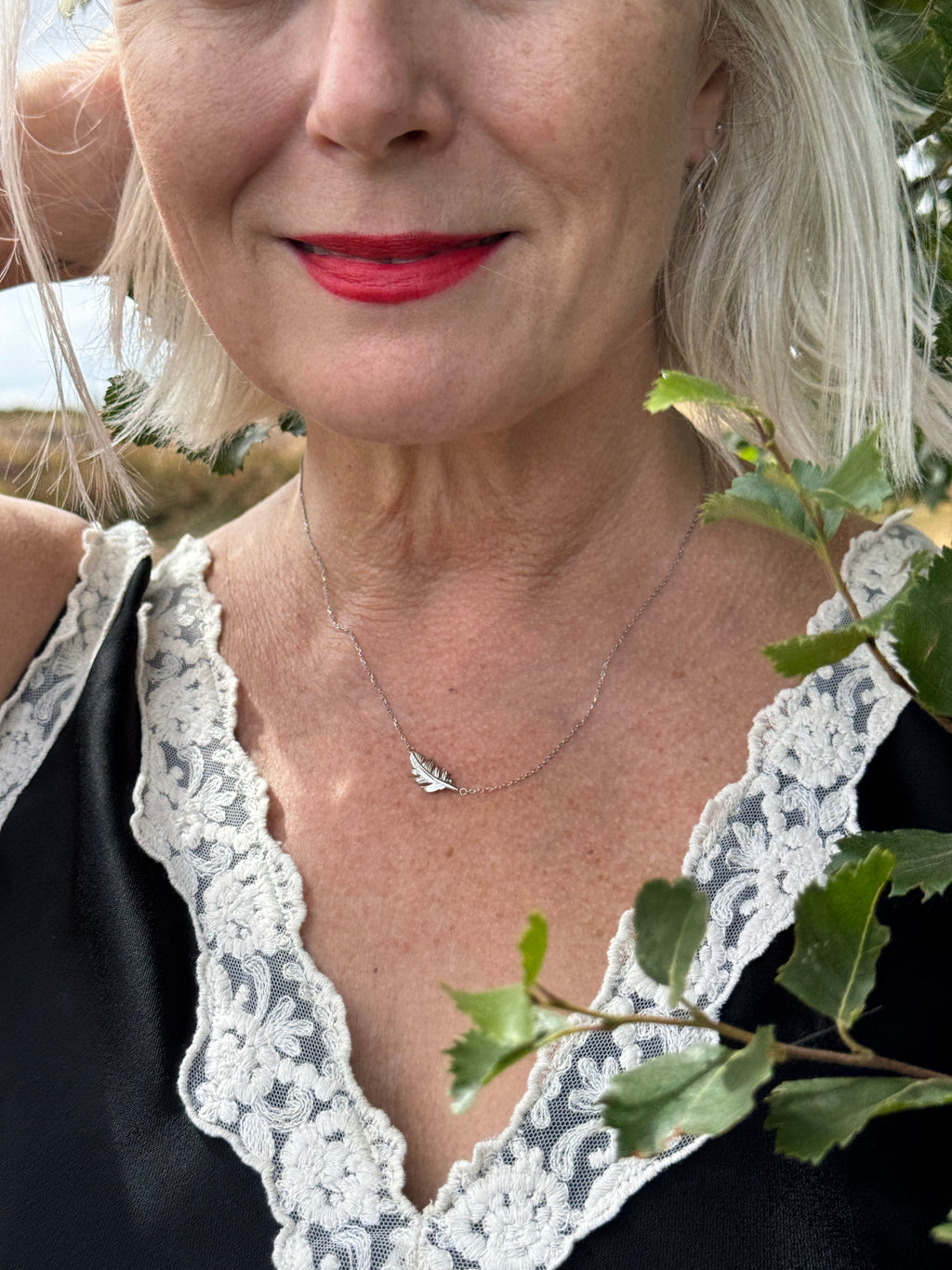 A close-up lifestyle shot of a woman wearing a delicate silver necklace with a small feather pendant, paired with a black top featuring white lace trim and green leaves hanging in the foreground.