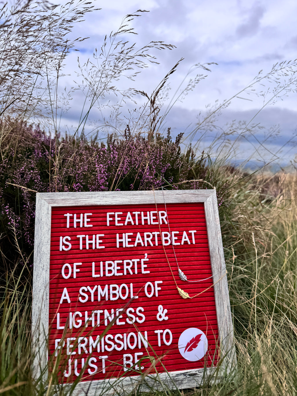 A red letter board sign with white text reading 'THE FEATHER IS THE HEARTBEAT OF LIBERTÉ A SYMBOL OF LIGHTNESS & PERMISSION TO JUST BE' placed outdoors in tall grass with purple heather and a cloudy sky in the background. A small feather pendant hangs from the sign.