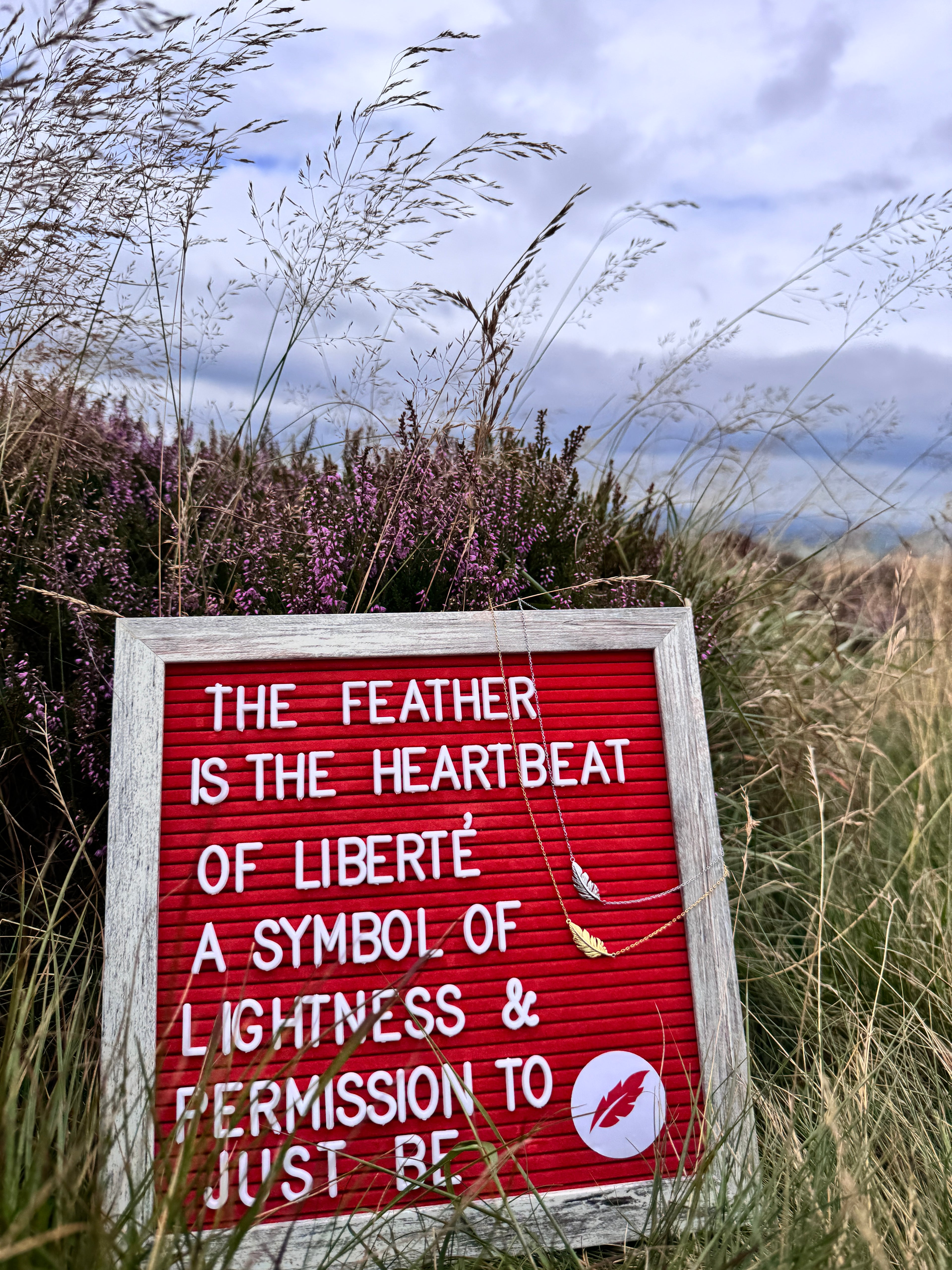 A red letter board sign with white text reading 'THE FEATHER IS THE HEARTBEAT OF LIBERTÉ A SYMBOL OF LIGHTNESS & PERMISSION TO JUST BE' placed outdoors in tall grass with purple heather and a cloudy sky in the background. A small feather pendant hangs from the sign.