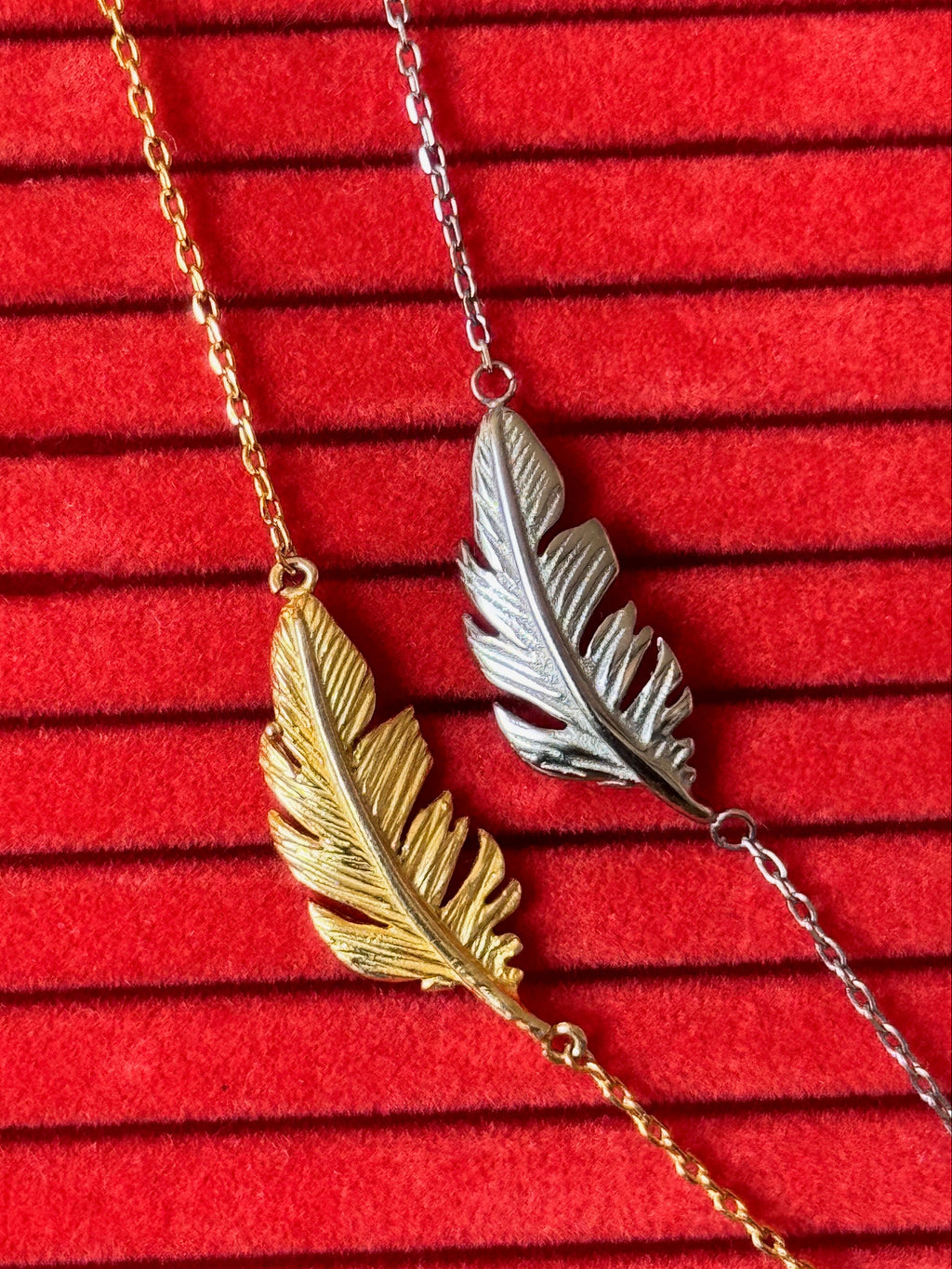 Two feather pendant necklaces, one in gold and one in silver, displayed on a red textured velvet background.
