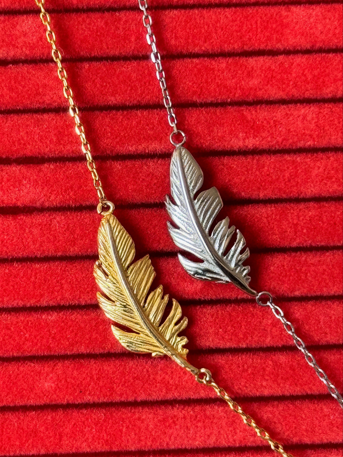 Two feather pendant necklaces, one in gold and one in silver, displayed on a red textured velvet background.