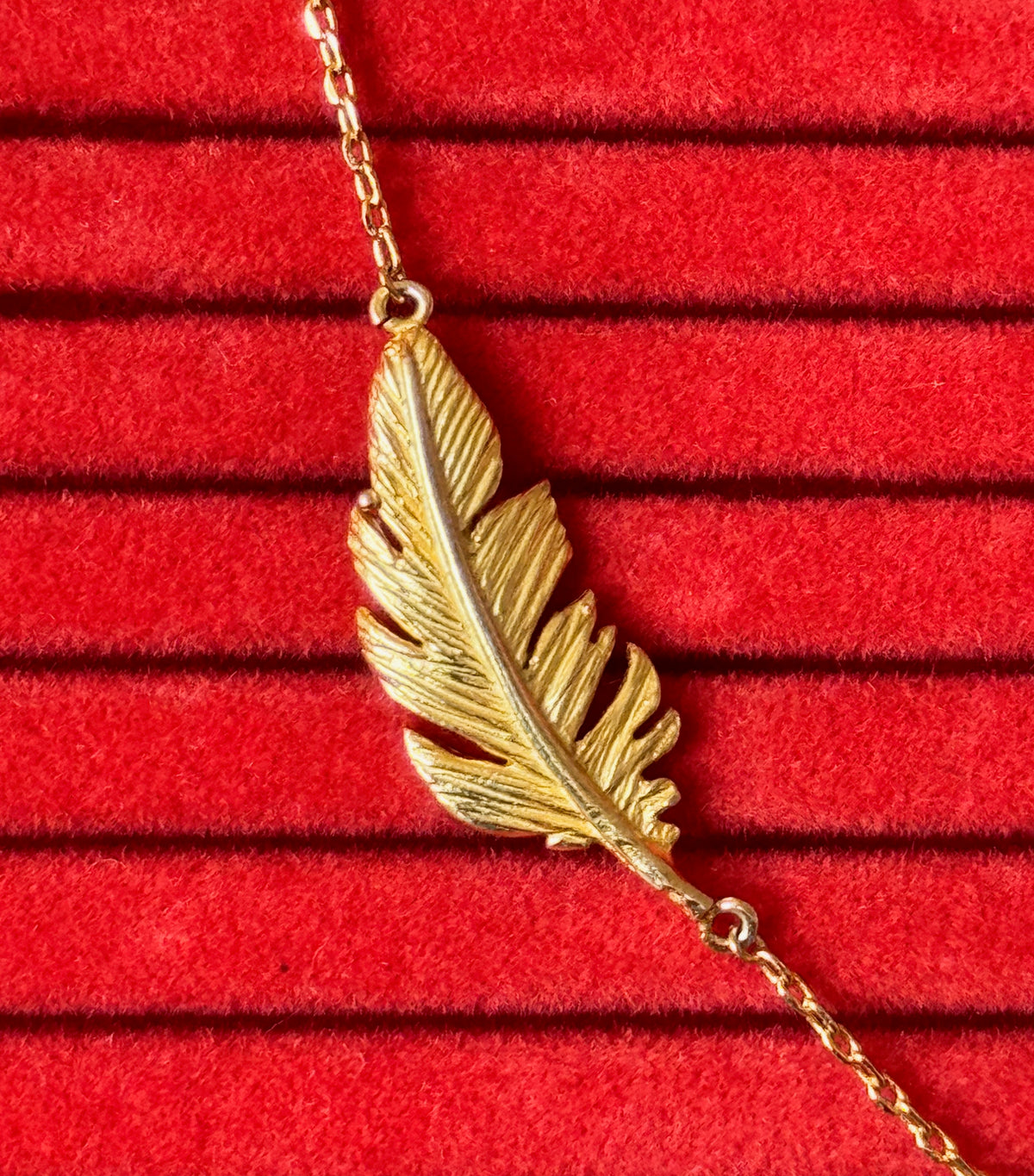 A gold feather pendant necklace on a matching chain, displayed on a textured red velvet jewelry box.