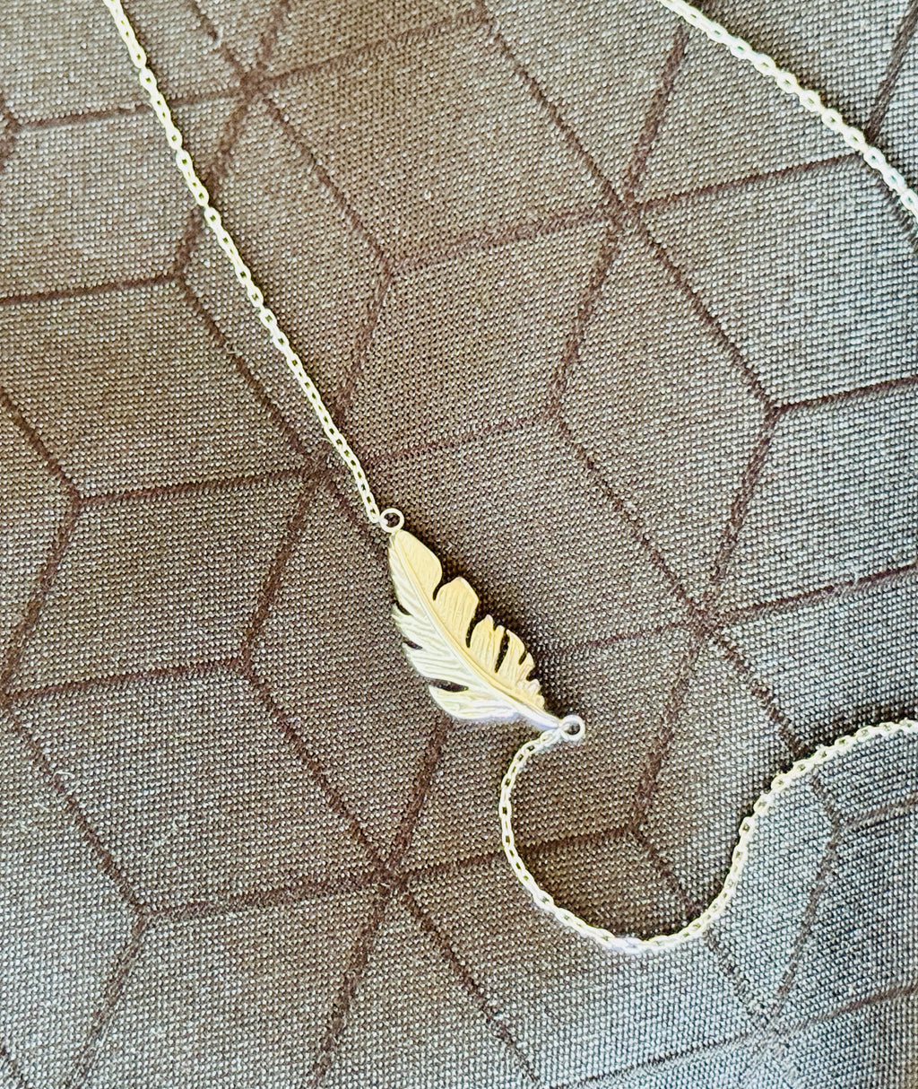 A silver feather pendant necklace on a silver-toned chain, laid flat on a textured grey geometric fabric background.