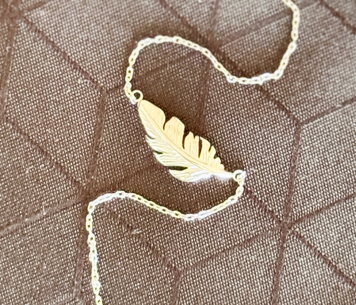 A gold feather pendant necklace on a delicate gold chain, displayed on a brown quilted fabric background.