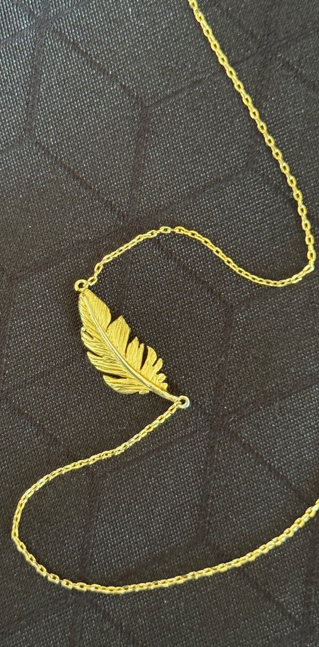 A gold feather pendant necklace on a delicate gold chain, displayed on a dark textured fabric background.