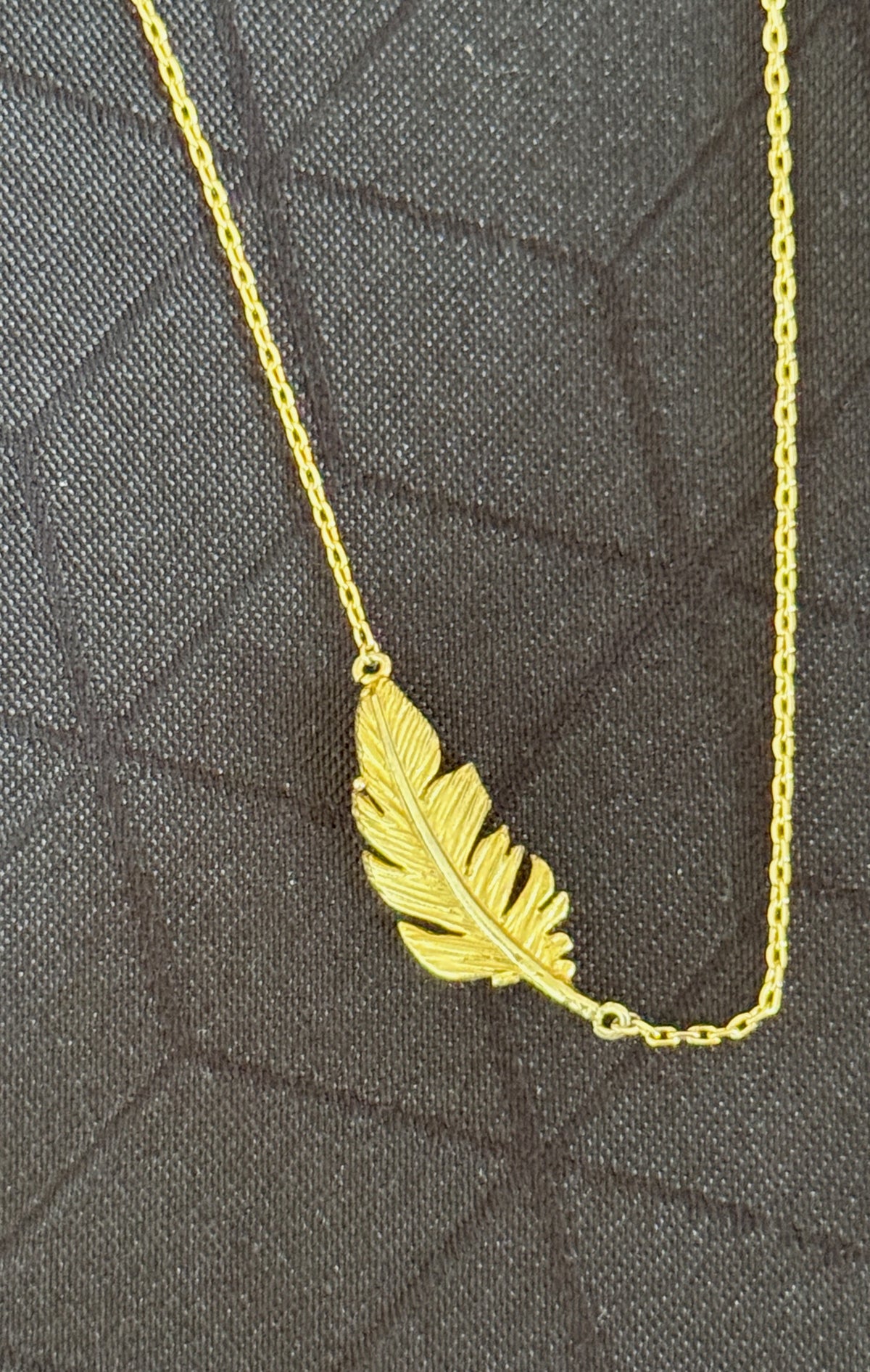 A gold feather pendant necklace on a matching chain, displayed on a dark textured fabric background.