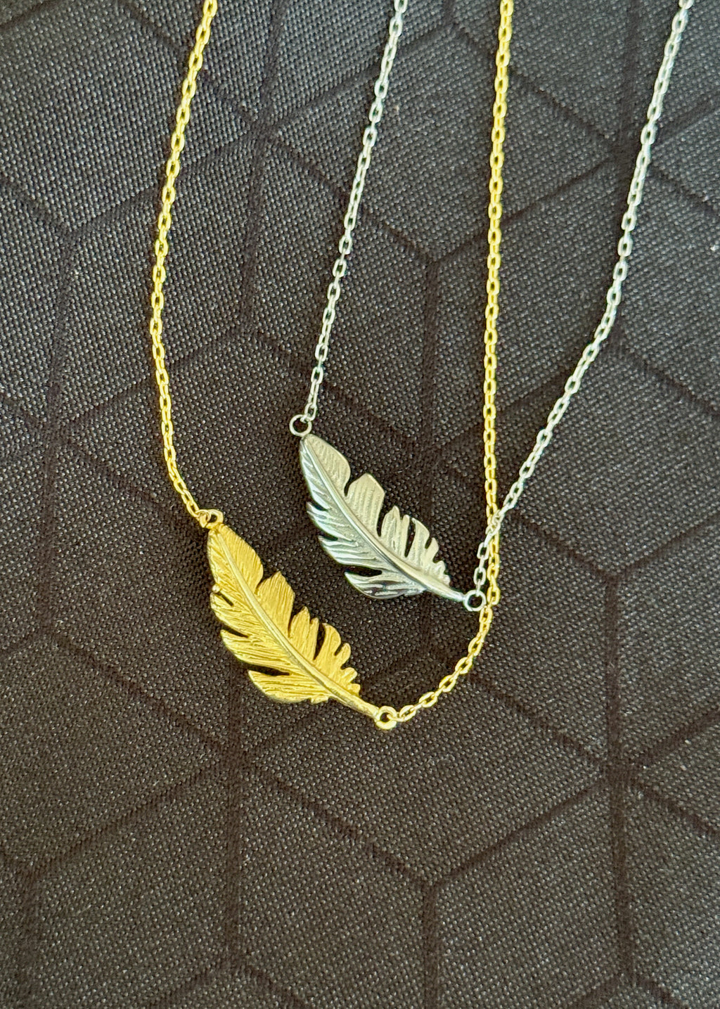 Two delicate chain necklaces with feather pendants, one in polished yellow gold and one in silver, displayed on a dark textured background.
