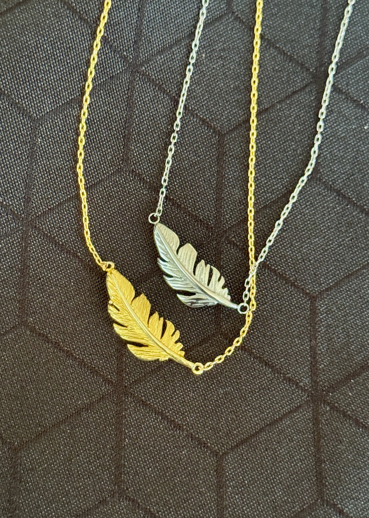 Two delicate chain necklaces with feather pendants, one in polished yellow gold and one in silver, displayed on a dark textured background.