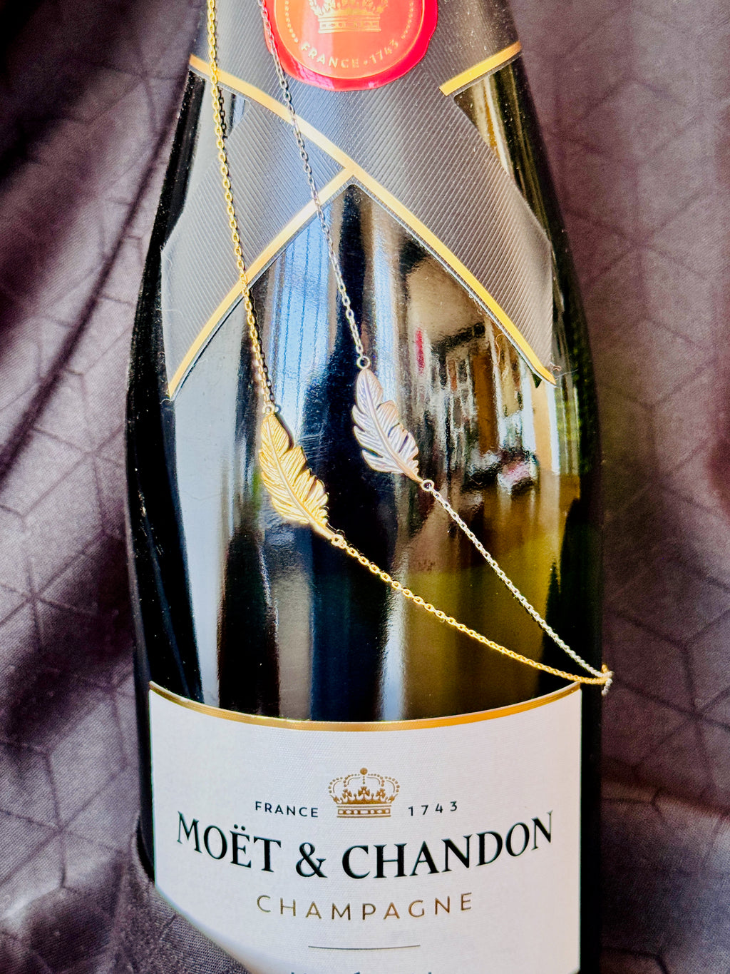 A gold feather pendant necklace draped over a Moët & Chandon champagne bottle, set against a dark textured fabric background.
