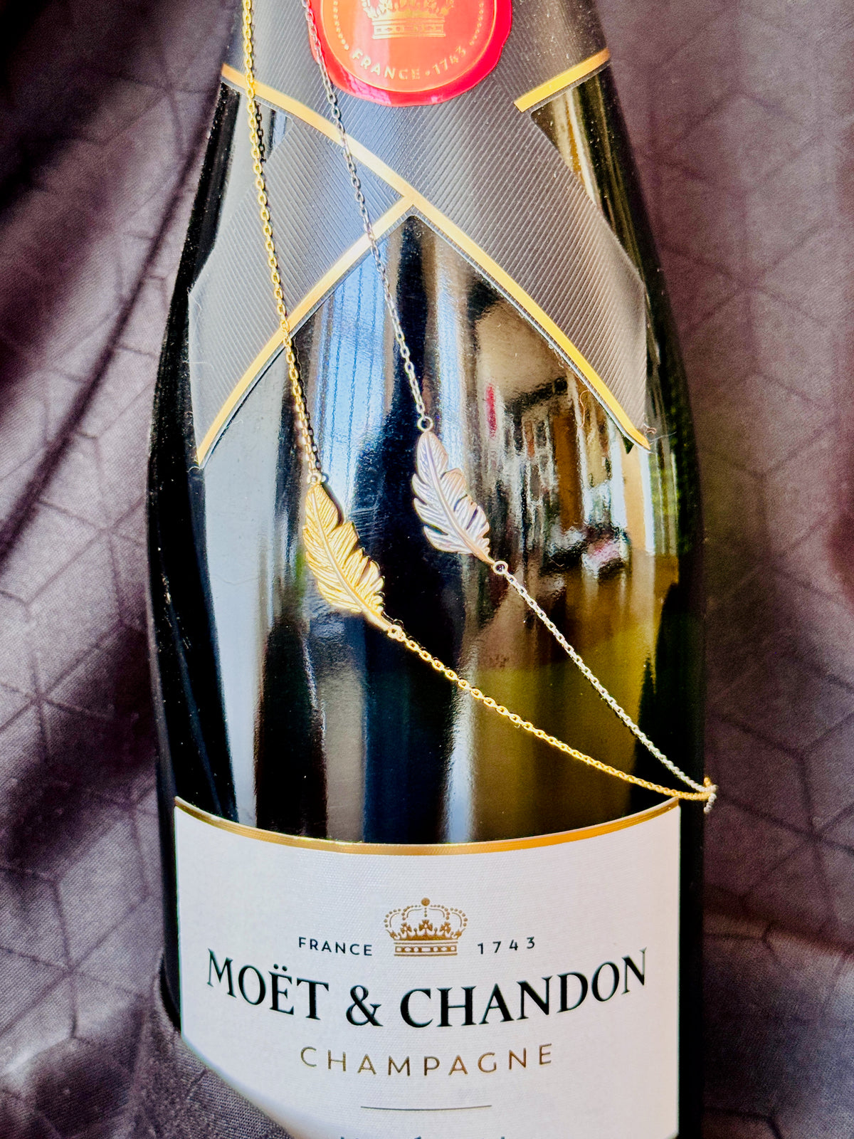 A gold feather pendant necklace draped over a Moët & Chandon champagne bottle, set against a dark textured fabric background.
