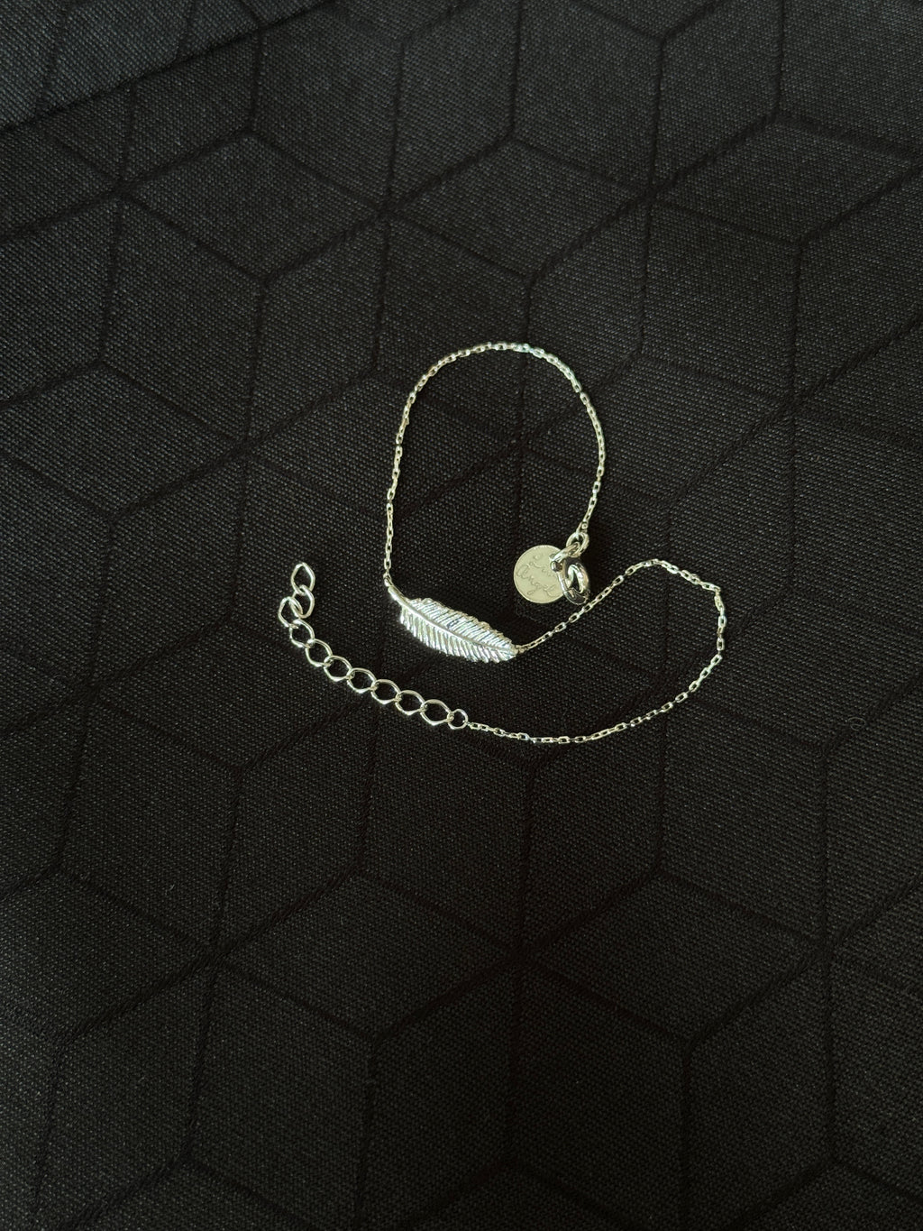 A delicate silver chain bracelet featuring a small feather charm and a round disc pendant, displayed on a textured black background.