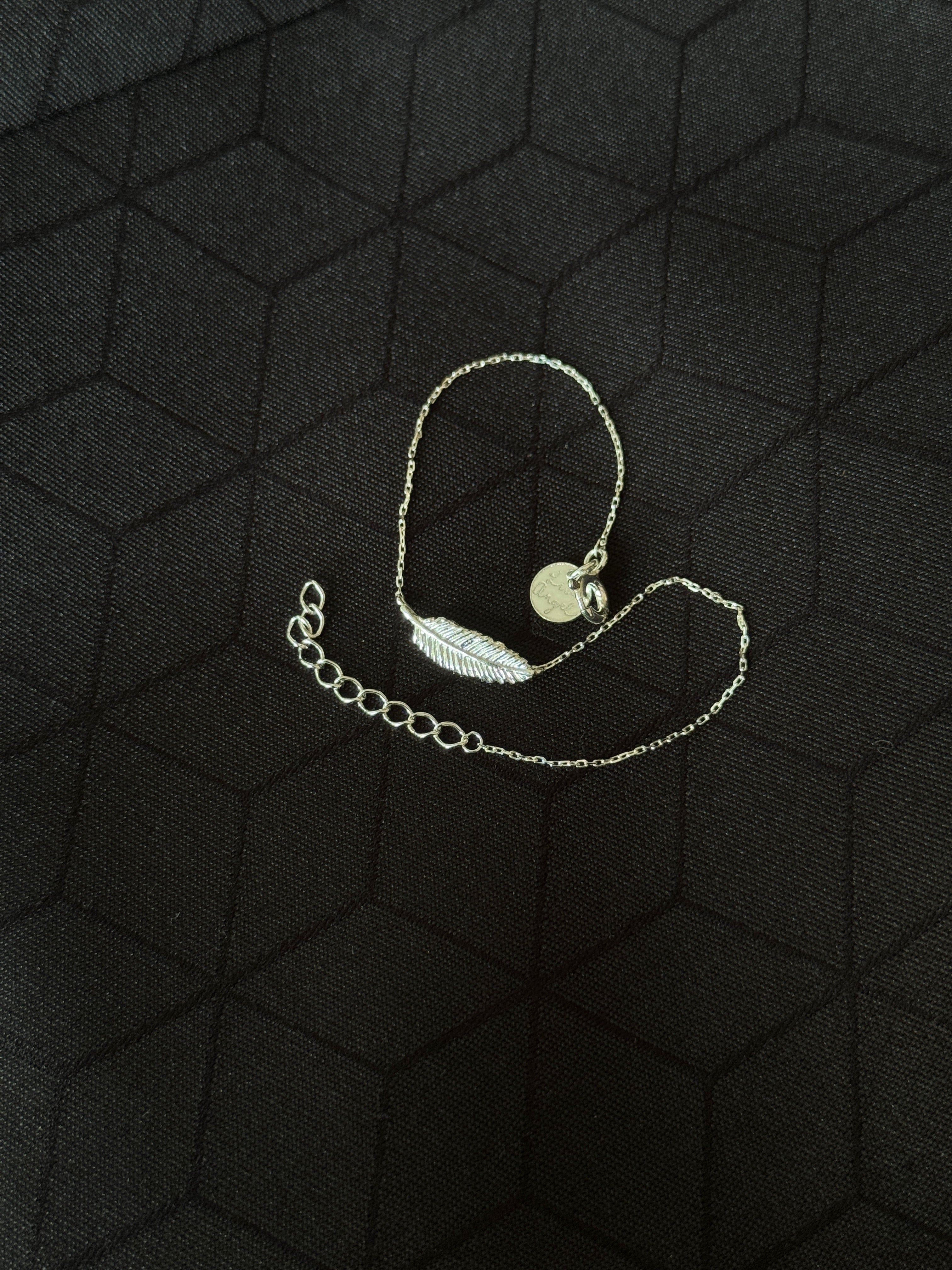 A delicate silver chain bracelet featuring a small feather charm and a round disc pendant, displayed on a textured black background.