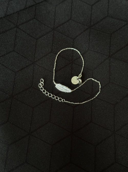 A delicate silver chain bracelet featuring a small feather charm and a round disc pendant, displayed on a textured black background.