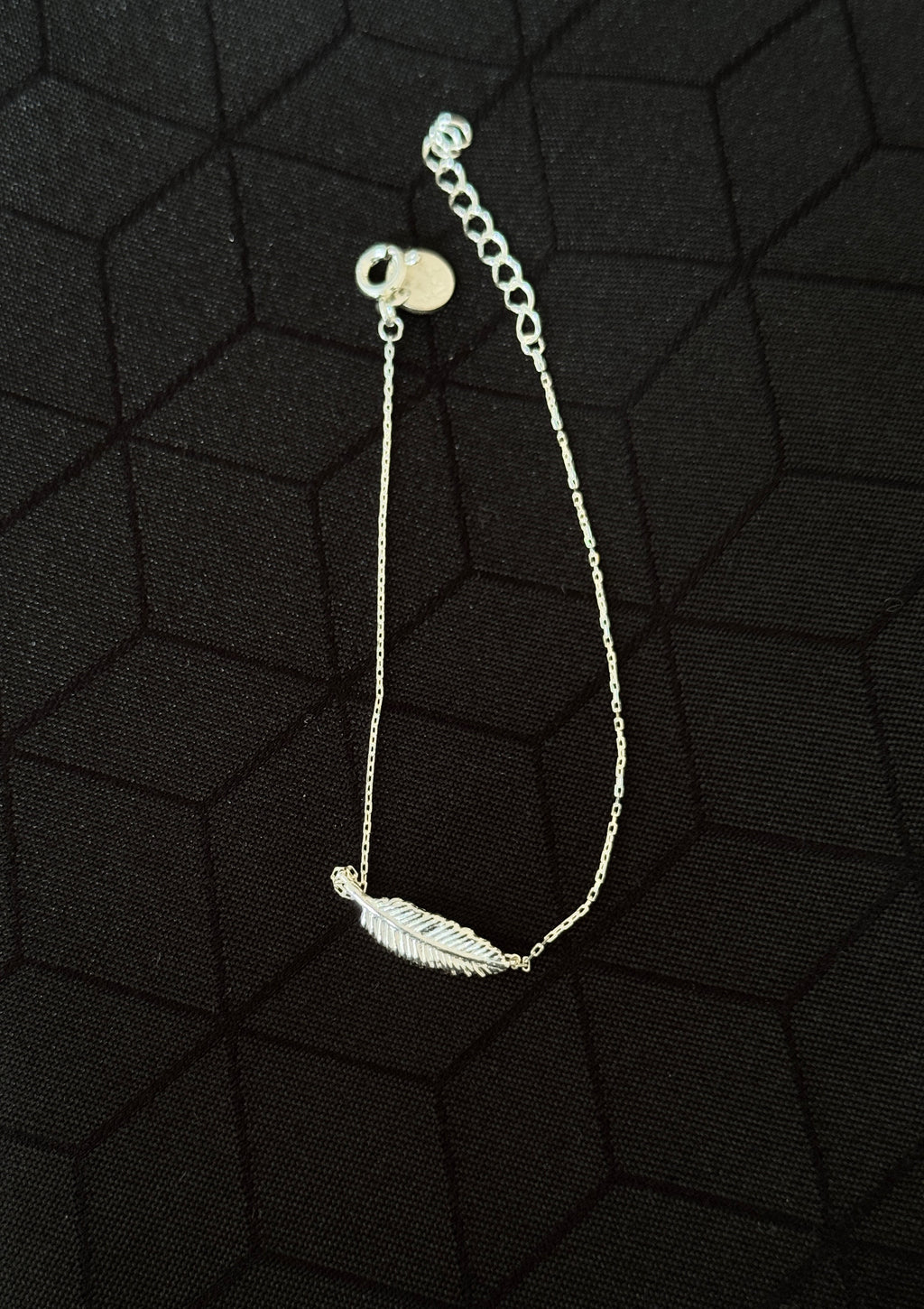 A delicate silver chain bracelet featuring a small feather charm, displayed on a textured black background.