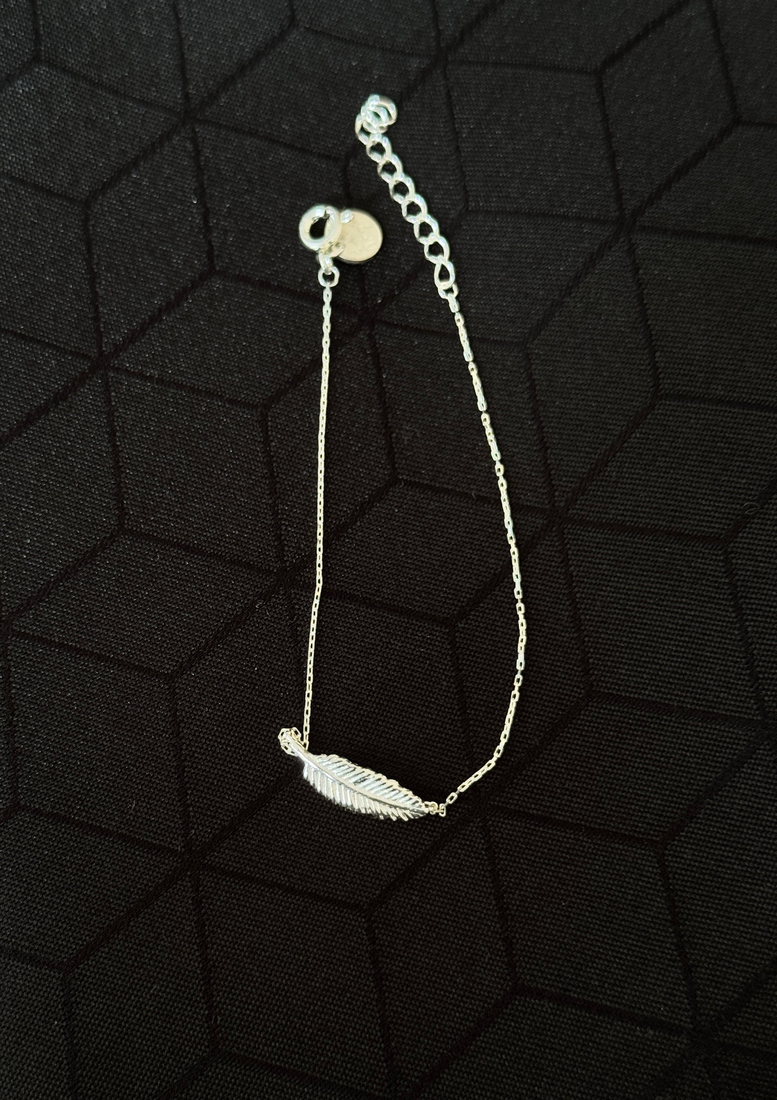 A delicate silver chain bracelet featuring a small feather charm, displayed on a textured black background.