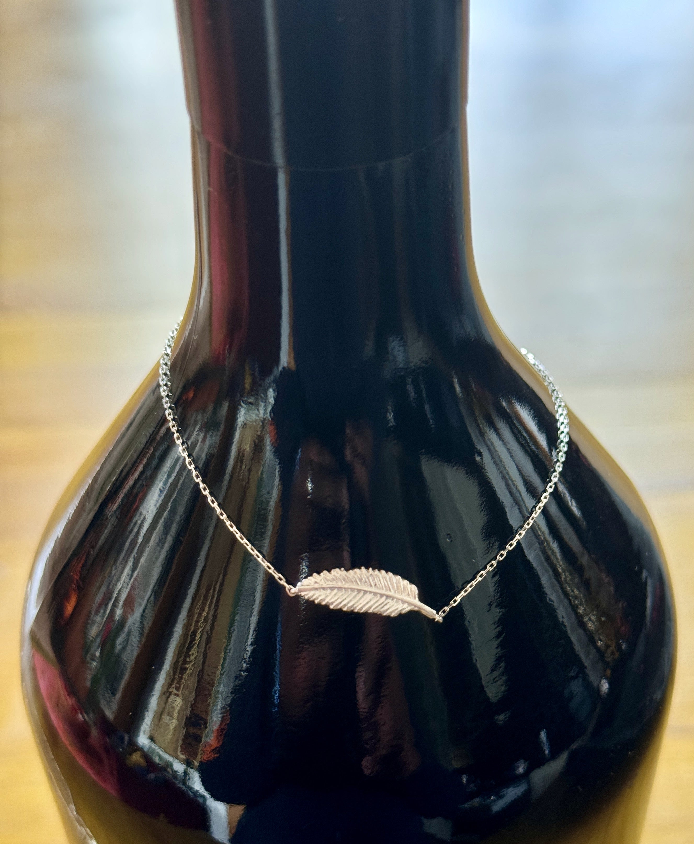 A delicate silver chain necklace with a small feather pendant, draped around the neck of a glossy black bottle against a softly blurred background.