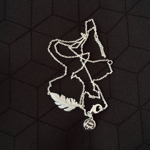A silver-toned necklace featuring a detailed feather pendant and a small round engraved charm, displayed on a black textured background.