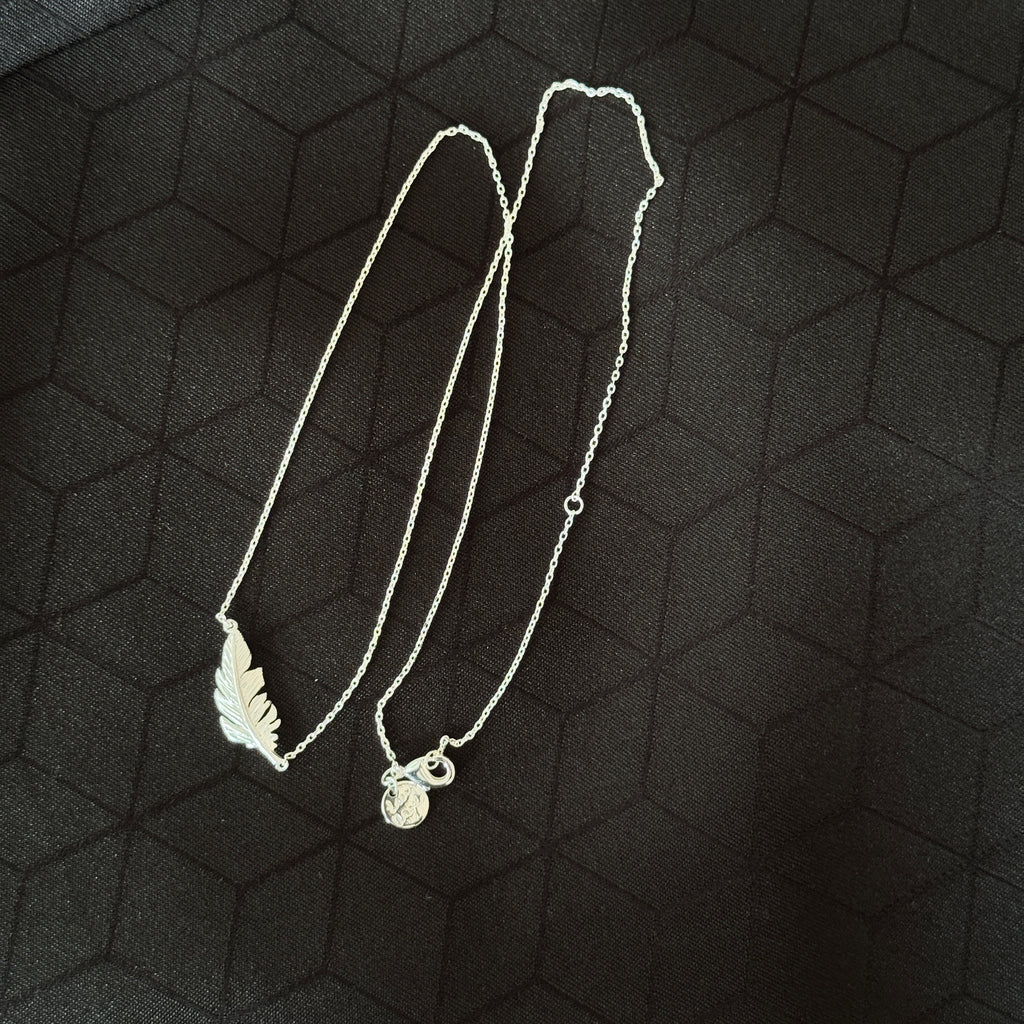 A silver necklace featuring a leaf-shaped pendant and a small circular charm, displayed on a dark textured background.