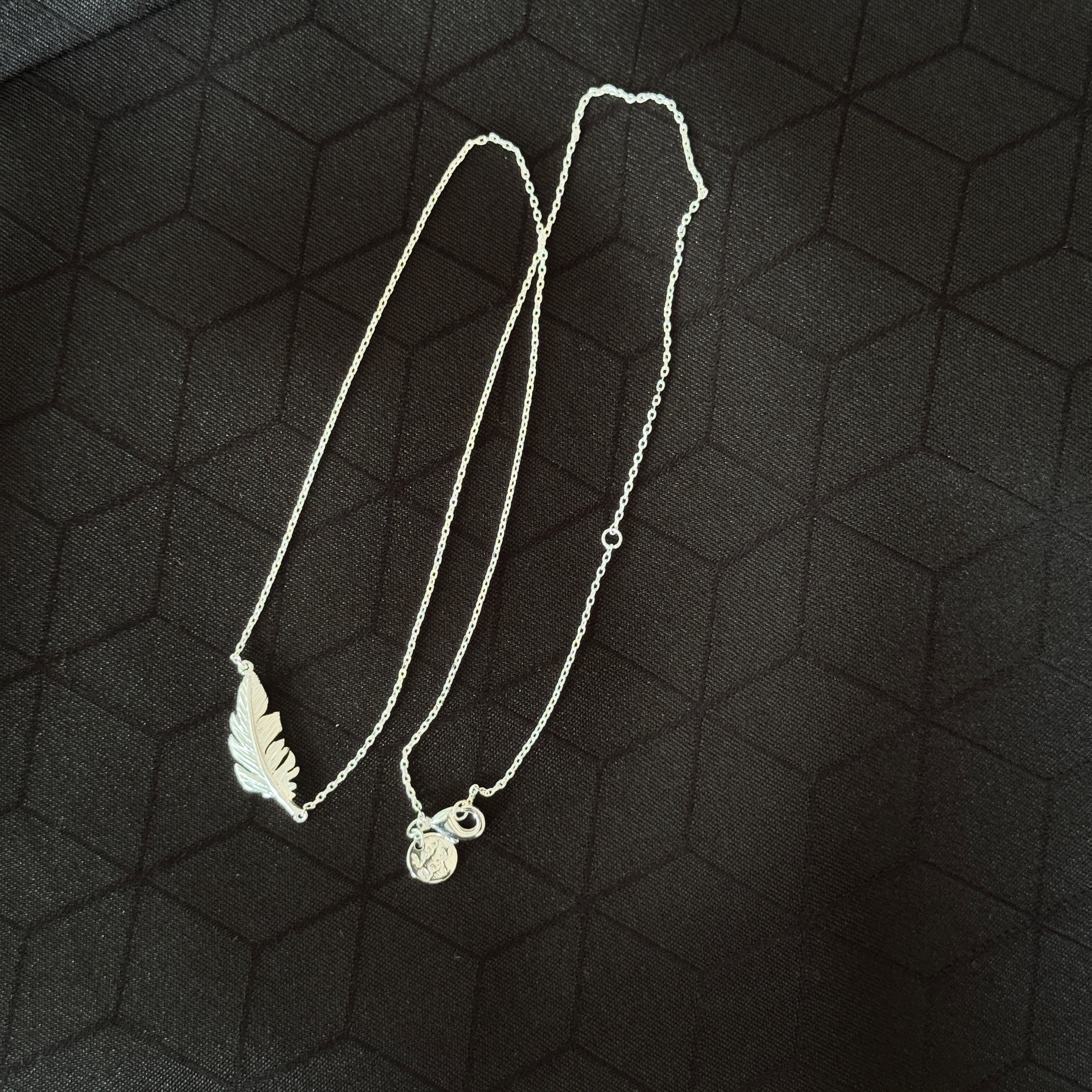 A silver necklace featuring a leaf-shaped pendant and a small circular charm, displayed on a dark textured background.