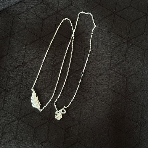 A silver necklace featuring a leaf-shaped pendant and a small circular charm, displayed on a dark textured background.