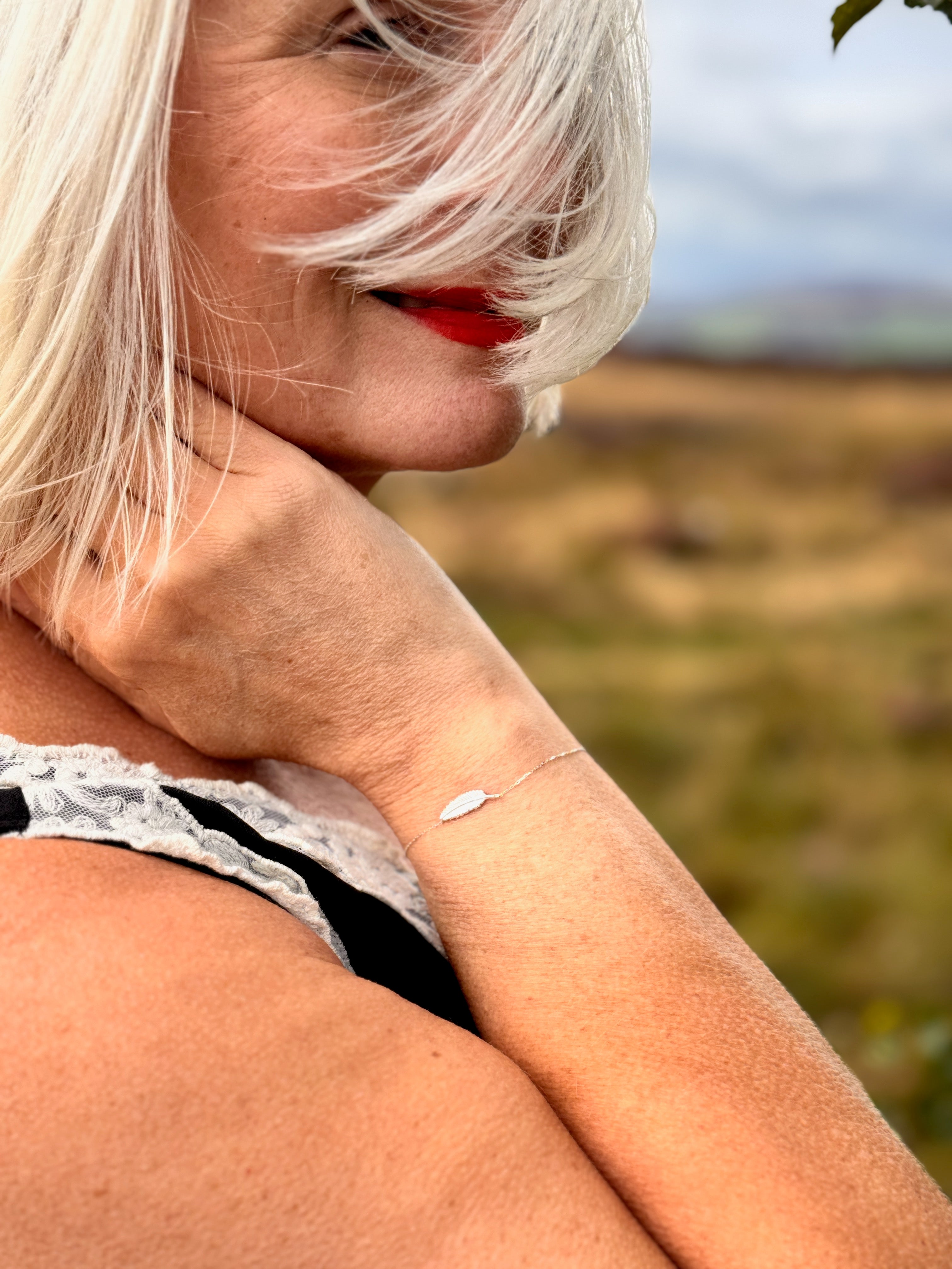 A close-up lifestyle shot of a woman with silver hair wearing a delicate silver leaf bracelet on her wrist, set against a blurred outdoor landscape.