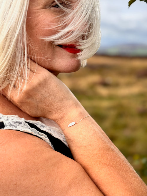 A close-up lifestyle shot of a woman with silver hair wearing a delicate silver leaf bracelet on her wrist, set against a blurred outdoor landscape.