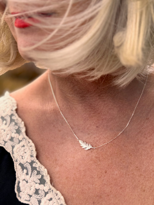 A close-up lifestyle shot of a woman wearing a delicate silver feather necklace. The image features a soft-focus background with warm, blurred tones, and the model is wearing a black top with a cream lace collar.