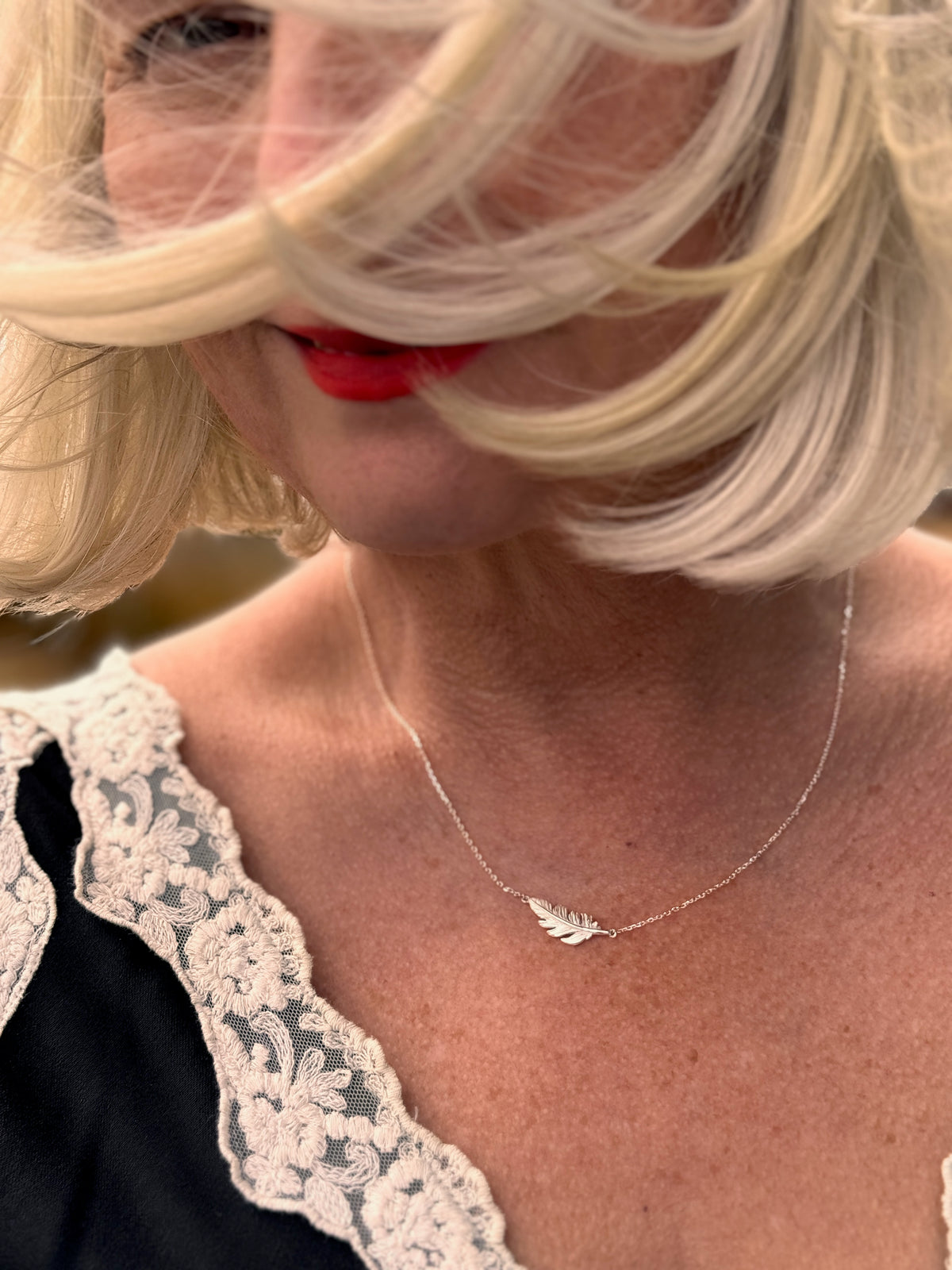 A close-up lifestyle shot of a woman with blonde hair blowing across her face, wearing a delicate silver necklace with a small leaf pendant and a black top with a cream lace collar.